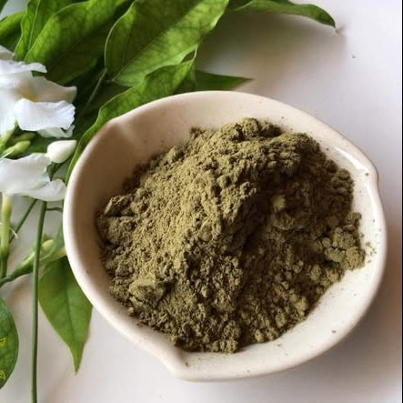 Bowl of green powder next to white flowers and green leaves on a light background
